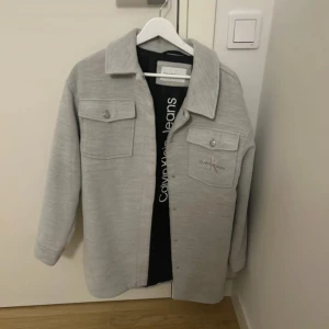 Ljusgrå overshirt från Calvin Klein Jeans - Snygg ljusgrå overshirt från Calvin Klein Jeans med klassisk krage, två bröstfickor med knappar och broderad logga på ena fickan. Jackan har raka ärmar och stängs med tryckknappar framtill. Perfekt för lager-på-lager-stil.