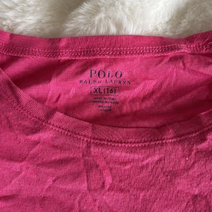 Rosa t-shirt från Polo Ralph Lauren - Säljer en rosa t-shirt från Polo Ralph Lauren i bomull. Modellen är klassisk med rund halsringning och korta ärmar. På bröstet finns den ikoniska blå broderade loggan. Perfekt för en chill och stilren look.