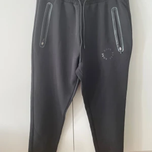 Svarta mjukisbyxor från BOSS - Svarta mjukisbyxor från BOSS med dragsko i midjan och två fickor med dragkedja. Diskret BOSS-logga tryckt på ena benet. Perfekta för chill eller träning, med en skön och avslappnad passform.