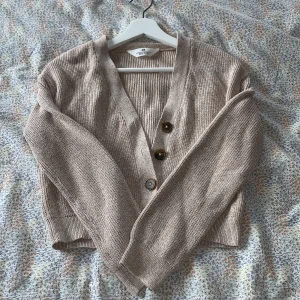 Beige ribbad kofta från H&M - Säljer en beige ribbad kofta från H&M i 100% bomull. Koftan har v-ringning, stora bruna knappar framtill och långa ärmar. Perfekt för lager-på-lager och enkel att matcha med jeans eller kjol.