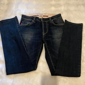 Mörkblåa jeans från Tramarossa - Snygga mörkblå jeans från Tramarossa med bootcut passform. Jeansen har klassiska fem fickor, kontrastsömmar och diskreta slitningar framtill. Materialet är robust denim med en skön känsla och detaljer i rött vid fickorna. Perfekta för en cool och avslappnad stil. Nytt pris 7000kr mitt pris 450kr