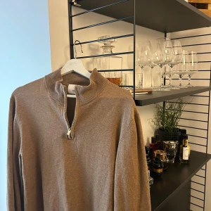 Beige stickad half zip-tröja från Bläck - Snygg beige stickad tröja från Bläck med half zip-dragkedja i halsen. Tröjan har en ribbad krage och långa ärmar, perfekt för lager på lager under höst och vinter. Enkel att matcha med jeans eller chinos för en clean look.