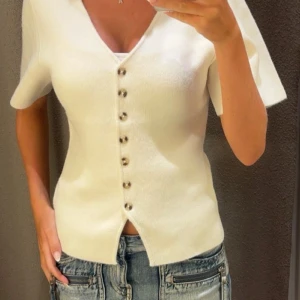 Vit v-ringad topp med knappar - Snygg vit topp med v-ringning och korta ärmar. Toppen har dekorativa knappar framtill och en liten slits nedtill. 