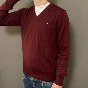 Vinröd v-ringad tröja från J.Lindeberg - Vinröd v-ringad tröja från J.Lindeberg med diskret logga på bröstet. Tröjan har långa ärmar, ribbade muddar och är gjord i mjuk bomull. Perfekt att bära över en t-shirt för en chill och stilren look.