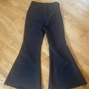 Svarta utsvängda byxor från Smiffys - Svarta bootcut byxor från Smiffys i 100% polyester. Byxorna stängs med knapp framtill. Vida ben med tydlig utsvängning ger en retro och cool look. 