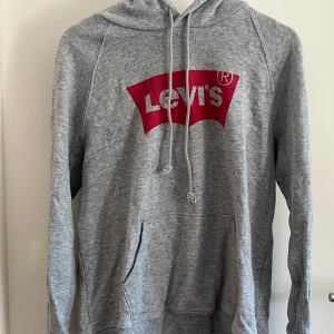 Grå hoodie från Levi's med röd logga - Säljer en grå hoodie från Levi's i storlek M. Tröjan har klassisk huva med snörning, magficka och en stor röd Levi's-logga på bröstet. 