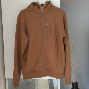 Brun hoodie från Sail Racing, M - Säljer en brun hoodie från Sail Racing i storlek M.  Använd max 2 gånger. Tröjan har hel dragkedja, huva med snörning och en bröstficka med dragkedja. Tillverkad i mjuk bomullsmix och har logga på bröstet och huvan. Perfekt för chill dagar.