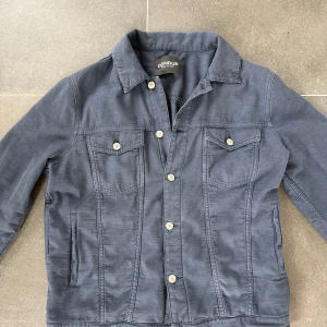 Mörkblå overshirt från Dondup - Snygg mörkblå overshirt från Dondup med klassisk krage och raka linjer. Overshirten har två bröstfickor med knappar, sidofickor och knäppning framtill. Perfekt nu för hösten, inte för varm och inte för kall. Storlek: 50/M