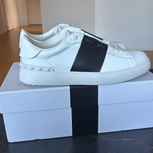 Valentino Garavani Open sneakers vit/svart - Säljer ett par Valentino Garavani Open sneakers i vitt skinn med bred svart rem över ovandelen och ikoniska nitar baktill. Klassisk rund tå, platt sula och vita snören. Snygg och stilren design som sticker ut med den svarta detaljen.