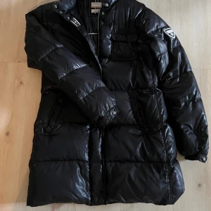 Svart pufferjacka från Champion XL - Säljer en svart, lång pufferjacka från Champion Outdoor Collection i storlek XL. Jackan har glansig finish, flera fickor med knappar och dragkedja framtill. Perfekt för kalla dagar med sin vadderade design och klassiska look. Champion-logga på ärmen och detaljer vid kragen.