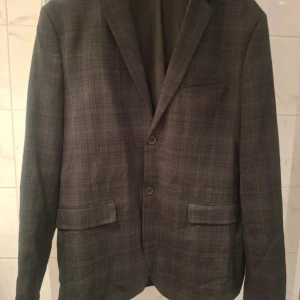 Grå / svart rutig kavaj från H&M Slim Fit - Snygg grå kavaj från H&M i slim fit-modell med diskret rutigt mönster. Kavajen har två knappar framtill, fyra knappar vid ärmslut och klassiska fickor med lock. Perfekt för dig som gillar stilrena detaljer och modern passform.