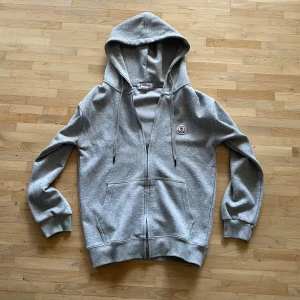 Moncler hoodie - Snygg grå hoodie från Moncler med dragkedja framtill, huva med snörning och klassisk logga på bröstet. Tröjan har ribbade muddar och två fickor framtill. Perfekt för en chill och stilren look.