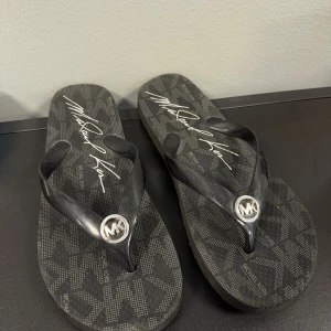 Svarta flip-flops från Michael Kors - Säljer nu mina fina svarta flip-flops från Michael Kors med mönstrad sula och tydlig logga i silver på remmen. Snyggt diskret mönster med MK-tryck och signatur på sulan💗💗