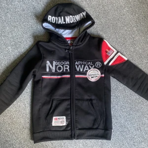 Svart hoodie Geographical Norway - (12years old är storleken)Svart hoodie från Geographical Norway med vit text på huvan och coola tryck på bröstet och ärmen. Röd och vit detalj på axeln, dragkedja framtill och två fickor. Perfekt för dig som gillar streetstyle och snygga loggor.