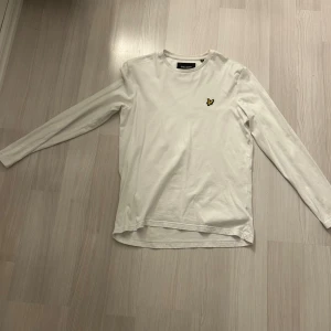 Vit långärmad tröja från Lyle & Scott - Vit långärmad tröja från Lyle & Scott med klassisk rund hals och den ikoniska gula loggan på bröstet. Tillverkad i mjuk bomull, perfekt för en clean och stilren look.