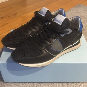 Svarta sneakers från Philippe Model - Snygga svarta sneakers från Philippe Model med blå och grå detaljer. Skorna har en klassisk design med snörning och en bekväm sula. Perfekta för en stilren look.