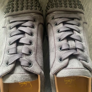 Christian Louboutin sneakers med nitar - Säljer ett par gråa sneakers från Christian Louboutin i mocka med ikoniska röda sulor och coola svarta nitar på tån. Skorna har snörning och rund tå, samt exklusiv känsla med märkets logga på innersulan. Perfekta för dig som vill sticka ut med lyxig streetstyle.