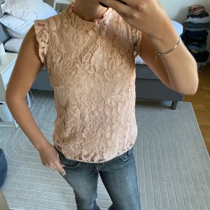 Rosa spetsblus med volangärm - Mer rosa i verkligheten Säljer en söt rosa blus i spets med blommigt mönster. Toppen har små volangärmar och hög hals med diskret volangkant. Perfekt att styla med jeans eller kjol för en romantisk look.