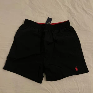 Svarta shorts från Polo Ralph Lauren - Svarta shorts från Polo Ralph Lauren med elastisk midja och röd broderad logga på ena benet. Klassisk och enkel design med bakficka och mjukt bomullsmaterial. Perfekta för sommardagar och chill vibes.
