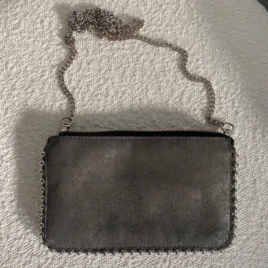 Svart axelväska med kedja och nitar - Cool svart axelväska i mocka med silverfärgad kedja som axelrem och snygga nitar längs kanterna. Väskan har två stora fack och ett mindre fack med dragkedja inuti, perfekt för att hålla ordning på dina grejer. Stängs med dragkedja upptill.