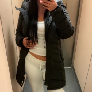 Svart lång dunjacka från Canada Goose - Säljer en svart lång dunjacka från Canada Goose med huva och dragkedja. Jackan har en quiltad design och klassisk patch på ärmen. Perfekt för kalla dagar och har en snygg, figurnära passform som håller dig varm hela vintern.