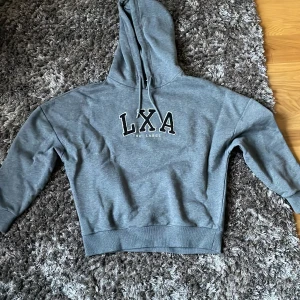 Grå hoodie från LXA The Label XS - Säljer en grå hoodie från LXA The Label i storlek XS. Tröjan har huva med snörning och tryckt logga framtill. Mjuk insida och ribbade muddar vid ärmslut och nederkant. Perfekt för chill dagar eller när du vill vara extra bekväm. Tröjan är helt ny!
