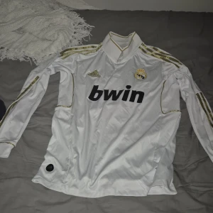 Real Madrid Ronaldo långärmad tröja - Säljer en officiell Real Madrid långärmad fotbollströja från Adidas med Ronaldo och nummer 7 på ryggen. Tröjan är vit med guldiga detaljer och Adidas-logga broderad på bröstet. Materialet är lätt och andas, Clima365-teknologi. Snygga guldiga ränder på axlarna.