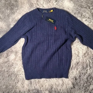 Marinblå stickad tröja från Polo Ralph Lauren - Snygg marinblå stickad tröja från Polo Ralph Lauren med ribbad struktur och klassisk röd logga broderad på bröstet. Rund hals och långa ärmar, perfekt för dig som gillar stilrena och tidlösa plagg.