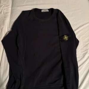 Svart sweatshirt från Stone Island - Svart sweatshirt från Stone Island med klassisk rund hals och ikonisk logotyp-patch på vänster ärm. Tröjan är i mjuk bomull och har ribbade muddar vid ärmslut och nederkant. Perfekt för dig som gillar streetwear och stilren design. Certilogo-verifierad