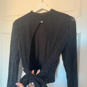 Glittrig svart topp med öppen rygg NA-KD - Supersnygg svart långärmad topp från NA-KD med glittriga silverränder. Toppen har öppen rygg och knytning i midjan, vilket ger en cool och trendig look. 