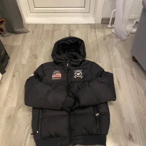 Svart pufferjacka Vinson Polo Club - Svart pufferjacka från Vinson Polo Club med huva, broderade detaljer och amerikansk flagga på bröstet. Jackan har dragkedja framtill, ribbade muddar och stor siffra 5 på ena ärmen. Perfekt för kalla dagar och har en sportig vibe.