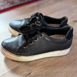 Svarta sneakers från Gant i skinn - Svarta sneakers från Gant med breda svarta snören och guldfärgade öljetter. Ovandel i skinn och vit sula som ger en clean kontrast. Klassisk rund tå och platt sula, perfekt för en chill och stilren look.