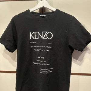 Svart Kenzo t-shirt med tryck - Svart t-shirt från Kenzo med vit text framtill och klassisk rund hals. T-shirten är kortärmad och har en clean, stilren look med tryck inspirerat av inbjudan till en modevisning. Perfekt för dig som gillar streetstyle och designerplagg.