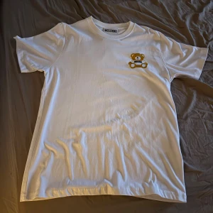 Vit Moschino t-shirt med nallebrodyr - Snygg vit t-shirt från Moschino med ett broderat nallemotiv på bröstet. Klassisk passform och rund halsringning. Perfekt statement-plagg för dig som gillar designer och lekfulla detaljer.