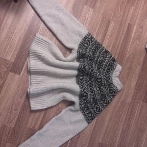 Grå stickad tröja från H&M - Mysig grå stickad tröja från H&M med svartmönstrad detalj över bröstet och axlarna. Tröjan har ribbade muddar och lång ärm, perfekt för kyliga dagar. Snygg och enkel att matcha till jeans eller kjol.