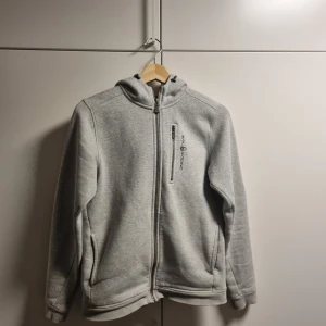Grå hoodie från Sail Racing - Snygg ljusgrå hoodie från Sail Racing med hel dragkedja och huva. Tröjan har en bröstficka med dragkedja och diskret logga på bröstet samt på huvan. Perfekt för kyliga dagar och enkel att matcha med det mesta.