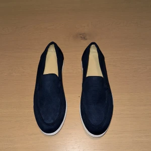 Mörkblå mockaloafers från ByCastello  - Snygga loafers i mörkblå mocka med stilren design och vit platt sula. Skorna har rund tå och är slip-on, vilket gör dem enkla att ta på. Insidan är ljusbeige och materialet känns mjukt och bekvämt. Perfekta för dig som gillar klassisk stil med modern touch. Orginalförpackning och dustbag medföljs.