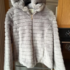Ljusgrå pälsjacka med huva Softy - Supermjuk ljusgrå pälsjacka från Softy outerwear med huva och horisontella ränder. Jackan har dragkedja framtill och är helfodrad för extra komfort. Perfekt för dig som vill ha en cozy och trendig look under kyliga dagar.