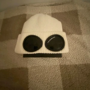 Cp company mössa - Snygg vit ribbad mössa med unika glasögondetaljer framtill. Perfekt för att sticka ut i mängden och hålla sig varm under kyliga dagar.ej original