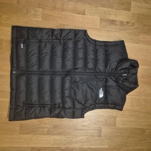 Svart dunväst från The North Face - Svart dunväst från The North Face med quiltad design och dragkedja framtill. Västen har en bröstficka med dragkedja och logga på bröstet, två sidofickor med dragkedja. Perfekt för lager-på-lager och håller dig varm under kyliga dagar. 