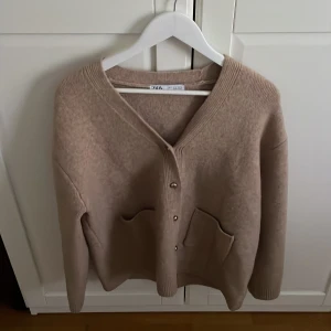 Beige kofta från Zara med fickor - Snygg beige kofta från Zara med guldfärgade knappar framtill. Koftan har två praktiska fickor och är tillverkad i ett mjukt stickat material som känns skönt mot huden. Perfekt för lager-på-lager och chill stil. DEN är helt oanvänd och köptes i slutet av sommaren i barcelona för 900kr. den passar till allt och är perfekt för vintern/hösten.