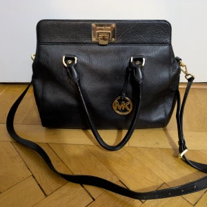 Svart handväska Michael Kors skinn - Stilren svart handväska från Michael Kors i äkta skinn med guldfärgade metalldetaljer och MK-logga. Väskan har både handtag och avtagbar axelrem, samt dragkedja och innerfack med logotypmönster. Perfekt storlek för allt du behöver och snygg att bära över axeln. Perfekt storlek och bra med fack. Köptes på Nordstrom i USA. 