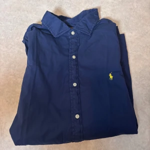 Blå skjorta från Polo Ralph Lauren XL - Snygg marinblå skjorta från Polo Ralph Lauren i bomull med klassisk krage och vita knappar. Custom fit-modell med gul broderad logga på bröstet. Perfekt för dig som gillar stilrena och tidlösa plagg med premiumkänsla.