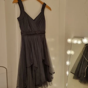 Svart tyllklänning med v-ringning - Supersnygg svart klänning med v-ringad överdel och breda axelband. Kjolen är i flera lager tyll som ger ett drömmigt och voluminöst intryck. Midjan markeras av ett brett band och klänningen har en elegant, asymmetrisk fåll.