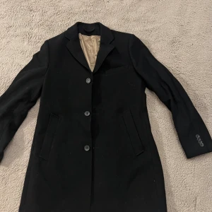 Svart kavaj från Bläck, strl 52 - Snygg svart kavaj från Bläck, modell Ashford Coat. Klassisk design med tre knappar framtill, två fickor och en bröstficka. Fodrad med beige insida och diskreta ränder. Perfekt för dig som gillar stilrena och tidlösa plagg.