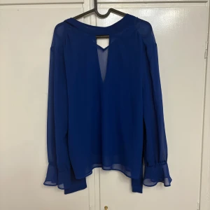 Blå blus med volangärm från Zara - Säljer en blå blus från Zara i transparent polyester med långa ärmar och volangdetaljer vid ärmslut. Blusen har v-ringning med knytband i halsen och en snygg öppen detalj framtill. Perfekt för dig som gillar stilrena men ändå unika plagg.