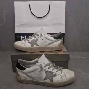 Golden Goose vita sneakers med stjärna - Golden Goose sneakers i vitt skinn med grå stjärndetalj på sidan och svart hälparti med logotyp. Skorna har grå snörning, rund tå och platt sula. Snyggt slitna för en cool streetstil. Perfekt för dig som gillar exklusiva sneakers med unik design.