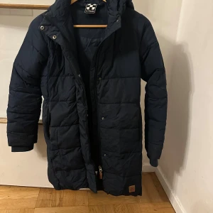 Mörkblå lång pufferjacka X-Trail - Säljer en mörkblå pufferjacka från X-Trail med huva och långa ärmar. Jackan har quiltad design, dragkedja och knappar framtill. Perfekt för kalla dagar med sin långa passform och snygga look. Fickor på sidorna och diskret logga nedtill.