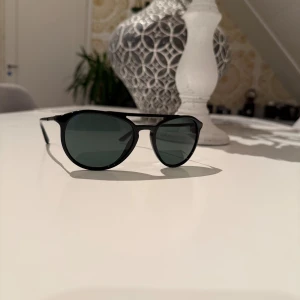 Svarta solglasögon Giorgio Armani - Snygga svarta solglasögon från Giorgio Armani med klassisk pilotform och mörka glas. Ramen är tunn och stilren, perfekt för dig som vill ha en tidlös och cool look. Diskret logga på skalmen ger en exklusiv känsla.