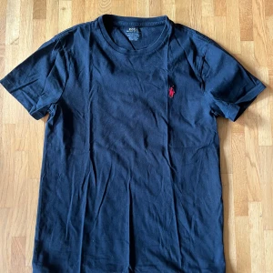 Svart t-shirt från Polo Ralph Lauren - Svart t-shirt från Polo Ralph Lauren med klassisk rund hals och röd broderad logga på bröstet. Modellen är custom slim fit och tillverkad i mjuk bomull som känns skön mot huden. Perfekt för dig som gillar stilrena och enkla plagg med en touch av lyx.